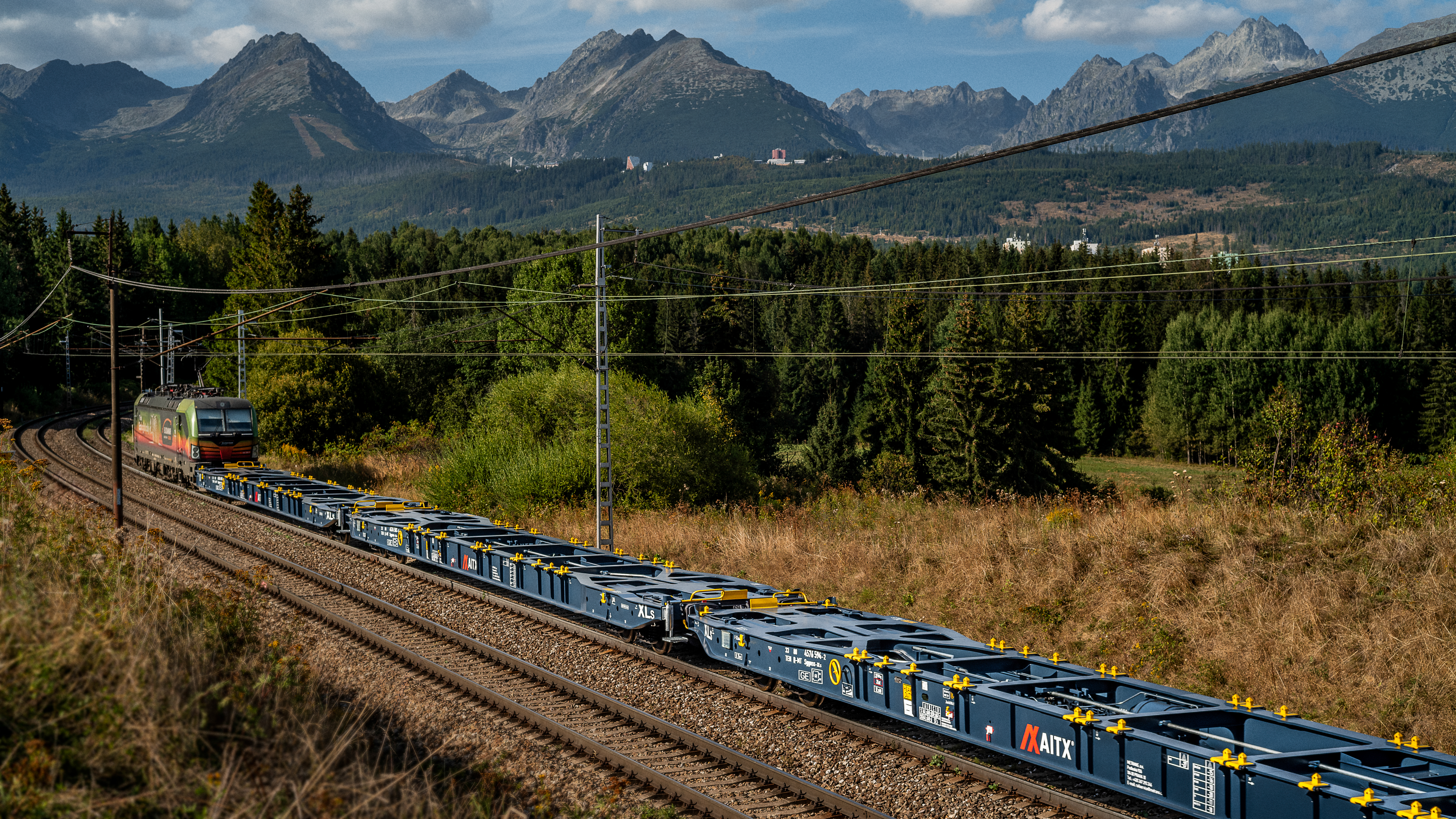 Train traveling through a scenic landscape in Europe, featuring a modern locomotive pulling flatbed railcars along a winding track, with majestic mountains and lush forests in the background.