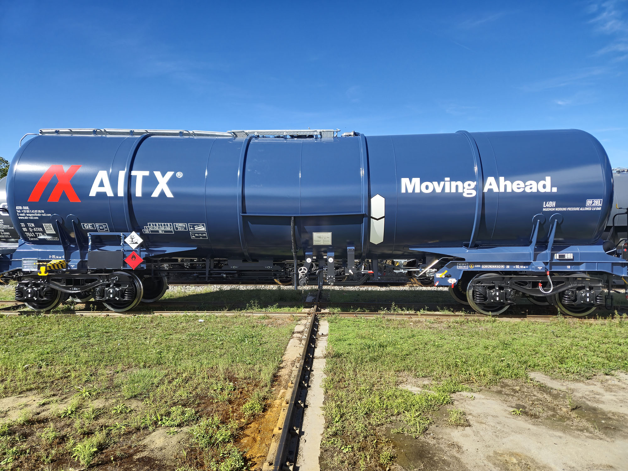 Blue freight train tank car labeled with "AITX" and "Moving Ahead" parked on tracks, surrounded by grass and clear blue sky, representing transportation and logistics in Europe.