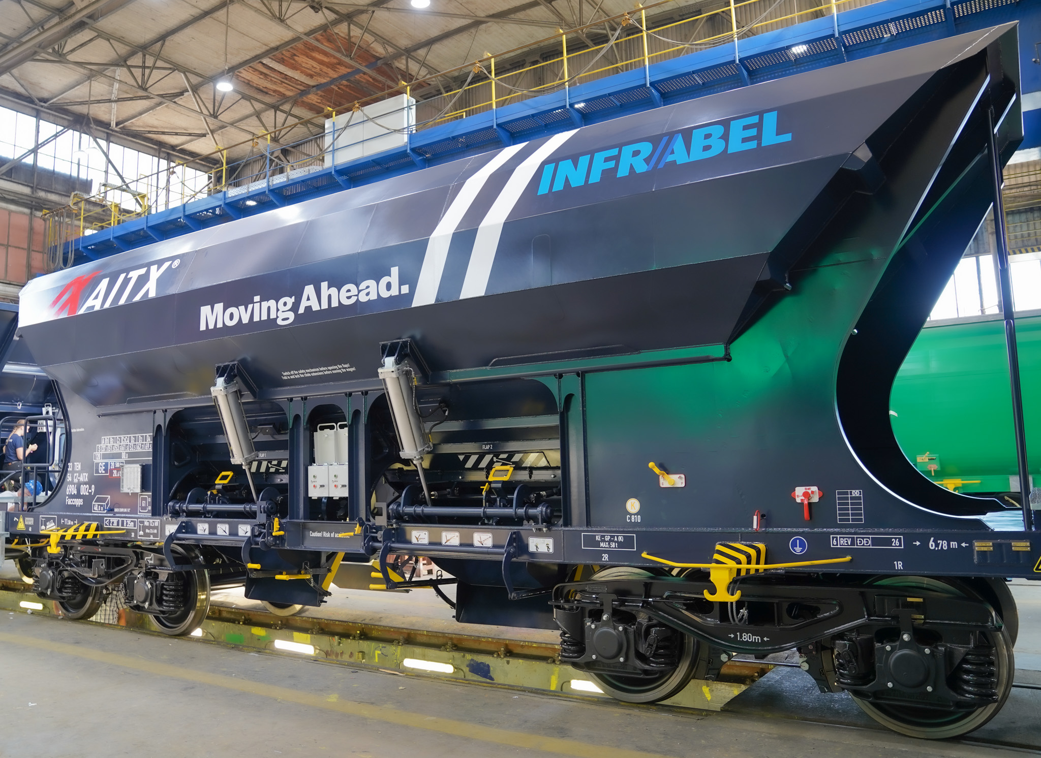Detailed view of a modern freight train car designed for transporting bulk materials, showcasing the branding of AITX and Infrabel, inside a well-lit industrial facility in Europe.