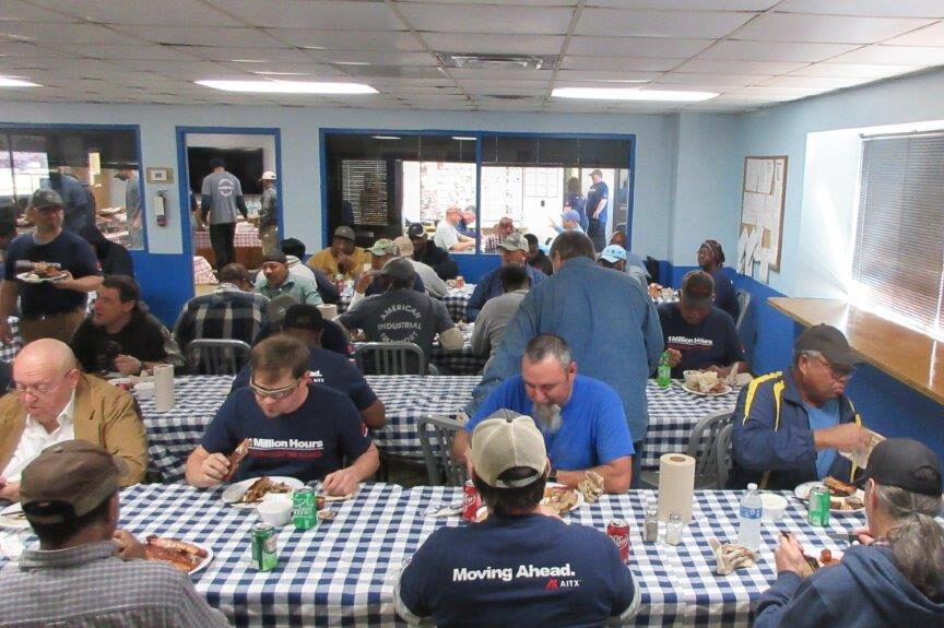 Group of workers enjoying a meal in a cafeteria setting, seated at tables with blue checkered tablecloths. The atmosphere is casual, with individuals engaged in conversation and eating various dishes. The room features large windows and a bright blue wall, creating a welcoming dining environment.