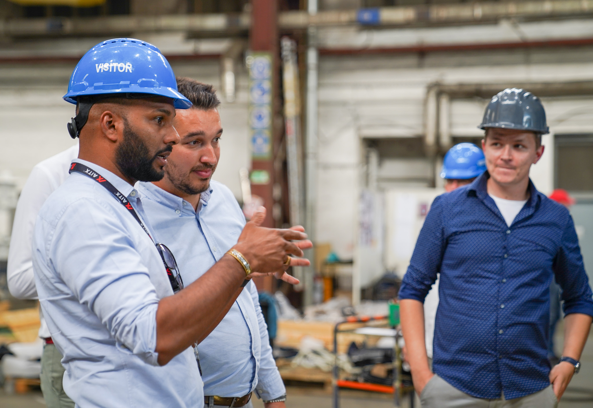 Group of professionals discussing in an industrial setting, wearing safety helmets, with one individual gesturing while explaining concepts related to manufacturing or engineering in Europe.