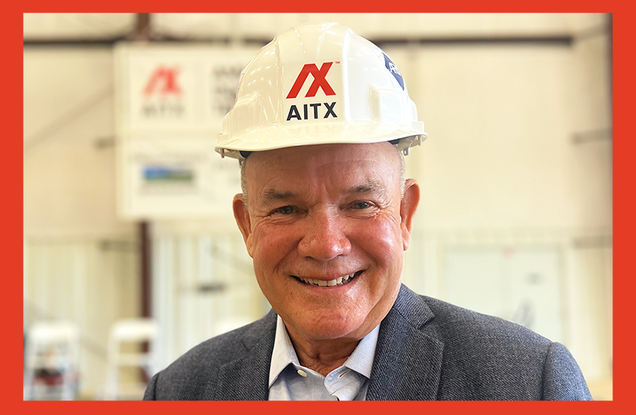Portrait of a smiling man wearing a hard hat with the AITX logo, standing in a construction or industrial setting, conveying professionalism and leadership in the construction industry.