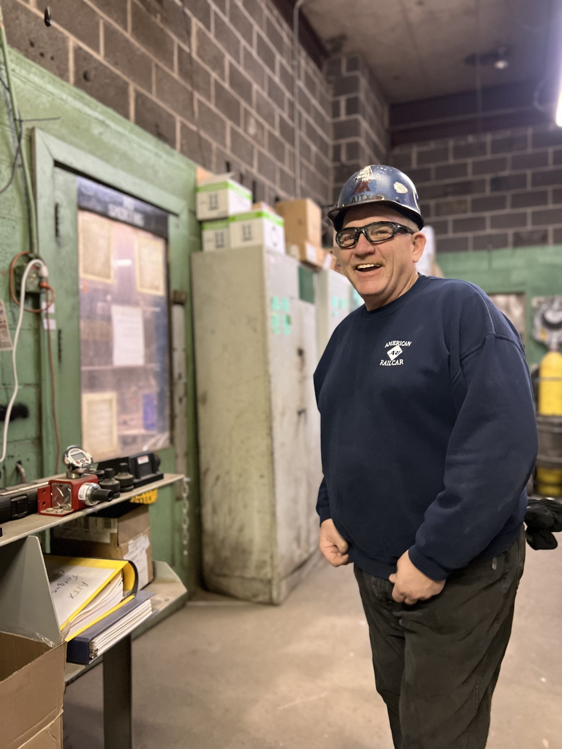 Smiling factory worker wearing a hard hat and safety glasses, standing in a workshop with industrial equipment and storage cabinets in the background.
