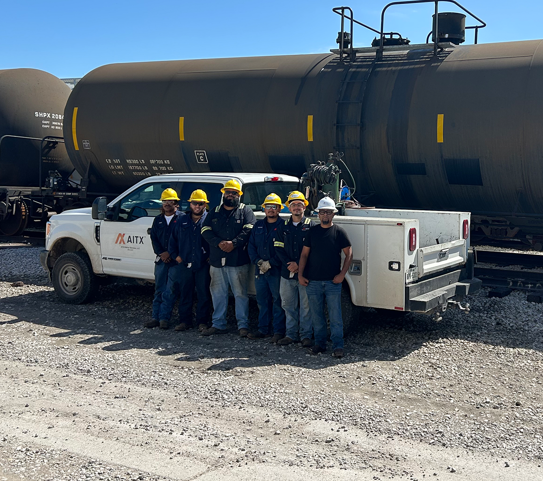 Group of workers in safety gear posing in front of a service truck and train tank cars, showcasing teamwork in an industrial setting.