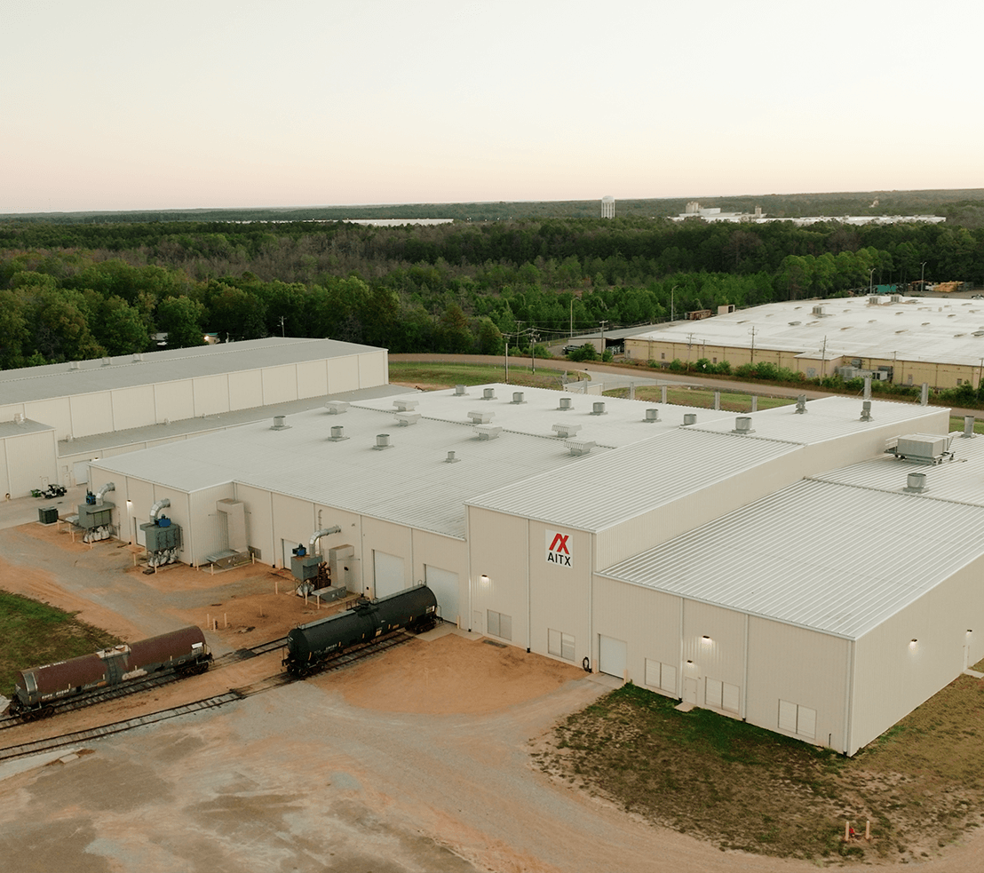 Aerial view of a large industrial facility with a metal roof, featuring the AITX logo, surrounded by greenery and a railway track with tank cars. The image captures the layout of the building and its operational environment.