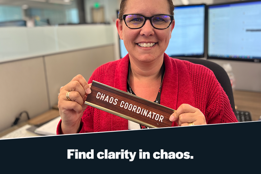 A smiling woman in a red cardigan holds a wooden sign that reads "CHAOS COORDINATOR" in an office setting with computer monitors in the background, illustrating a theme of finding clarity amid workplace chaos.