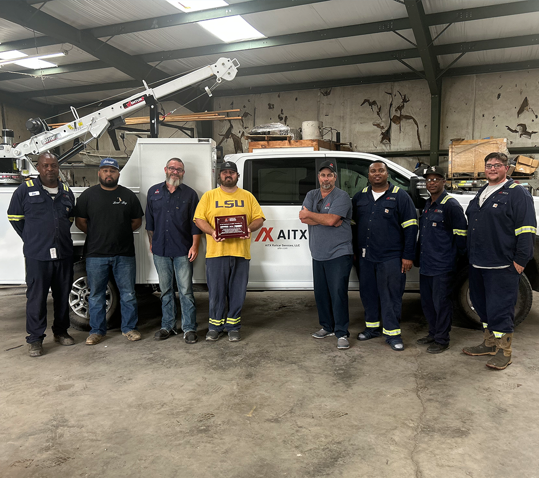 Team members of AITX Battery Services, LLC pose with an award in front of their work truck inside a warehouse. The group is dressed in work uniforms, showcasing teamwork and professionalism in the battery service industry.