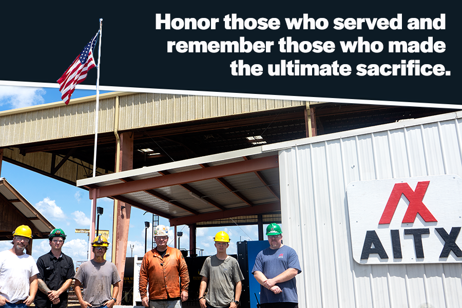 Group of workers in hard hats standing in front of a building with an American flag, promoting a message of honoring military service and remembering sacrifices made. The setting suggests a workplace, possibly in the construction or manufacturing industry.