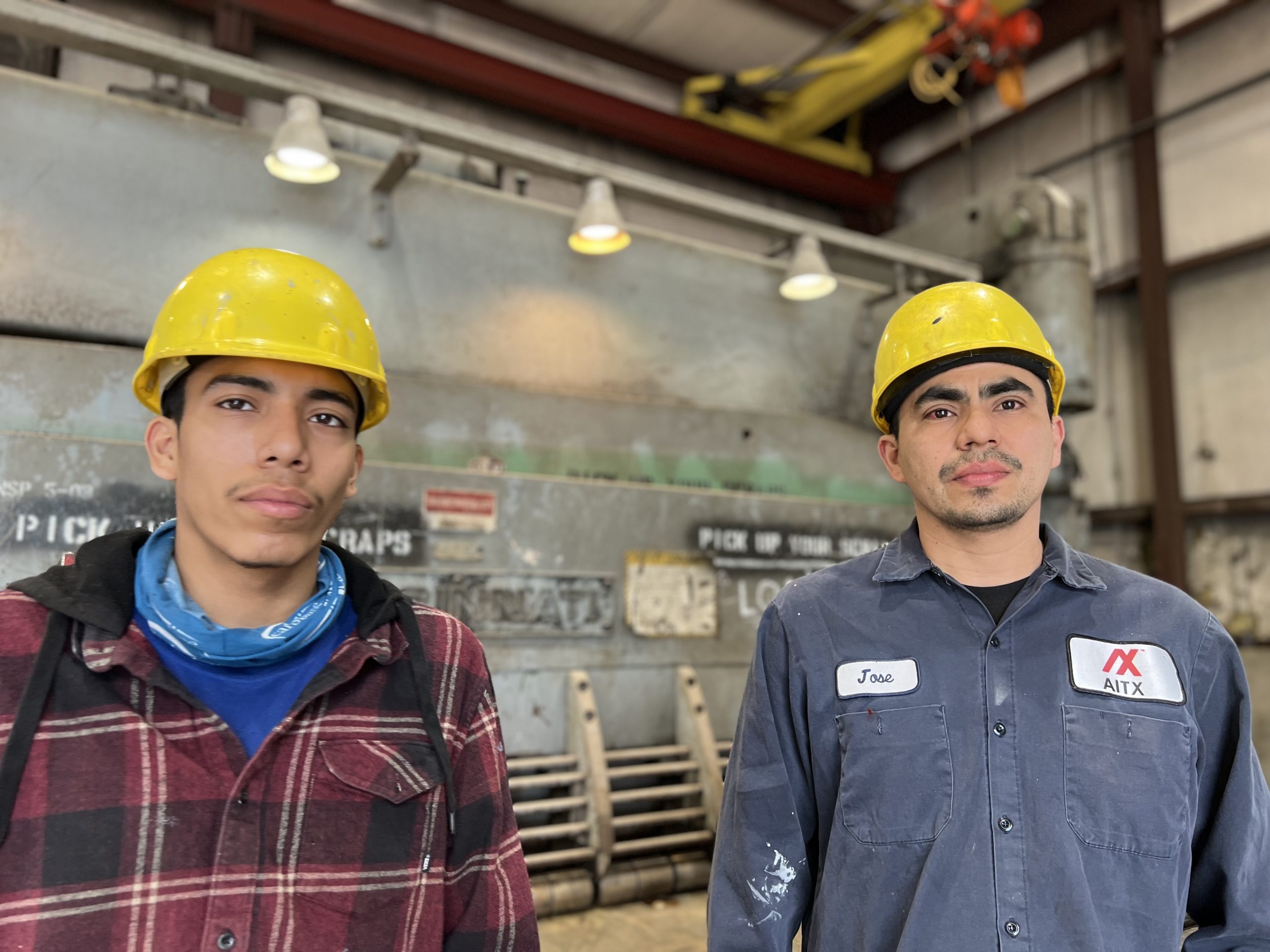 Two young men wearing yellow hard hats pose for a photo in an industrial setting. One is dressed in a plaid shirt and blue bandana, while the other wears a dark blue work shirt with a name tag. The background features machinery and signs, indicating a working environment focused on safety and teamwork.