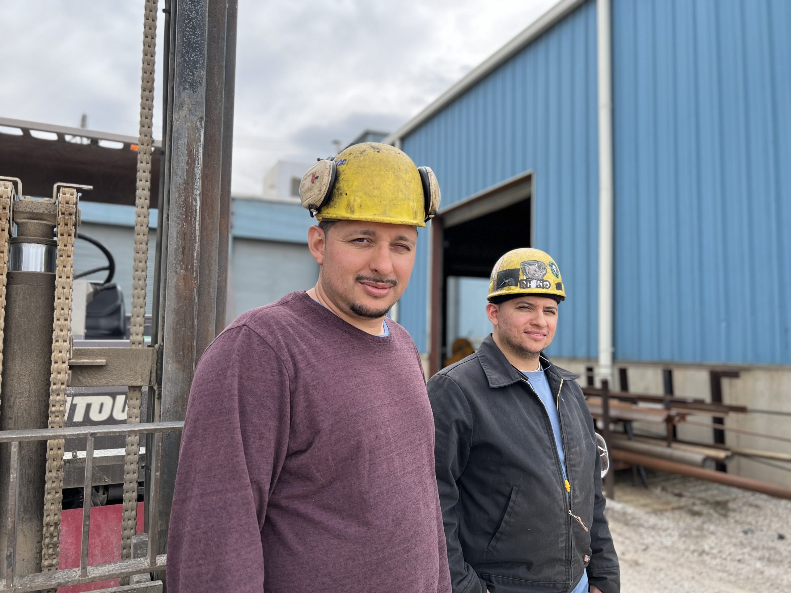 Two construction workers wearing safety helmets stand outside a blue industrial building, with a forklift visible in the background. The first worker wears a maroon sweater, while the second is dressed in a black jacket and blue shirt.