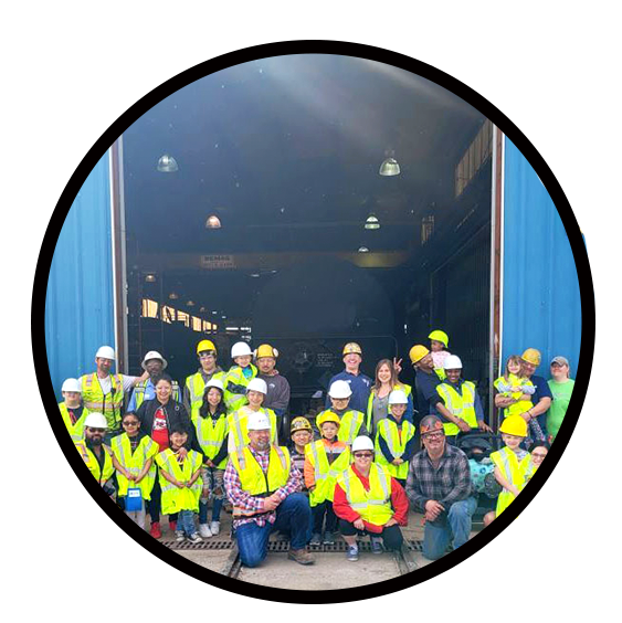 Group photo of diverse individuals in safety vests and hard hats, gathered in front of a large industrial building, showcasing teamwork and community engagement in a construction or safety event.