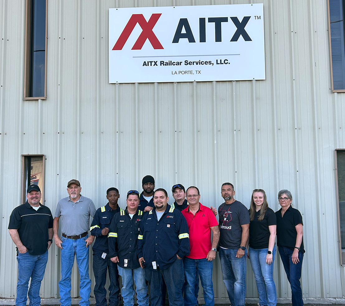 Group photo of AITX Railcar Services employees in La Porte, Texas, standing in front of the company sign. The team is composed of diverse individuals in work attire, showcasing a collaborative work environment in the rail services industry.