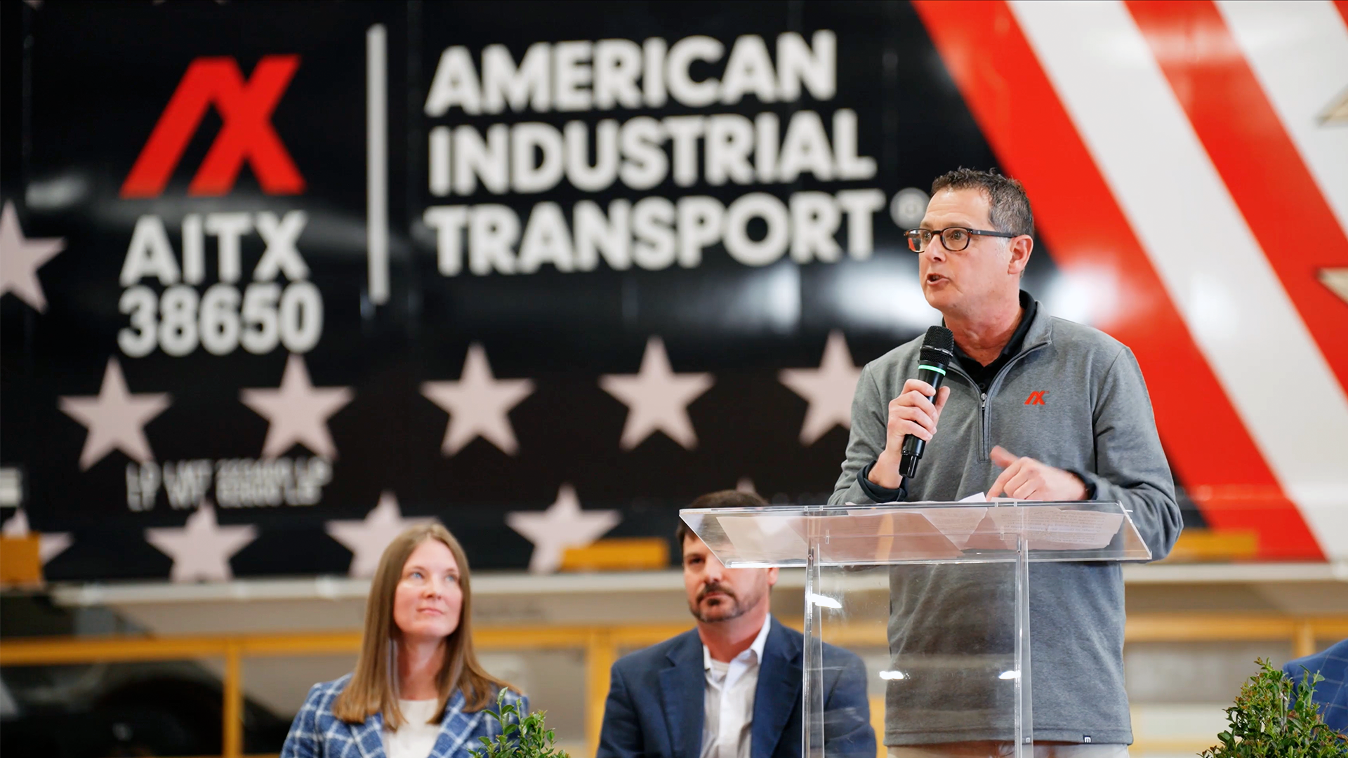 A speaker addresses an audience during an event for American Industrial Transport, with the company logo visible in the background. Two attendees listen attentively, highlighting engagement in the transportation industry. Peter Appel celebrates America 250.