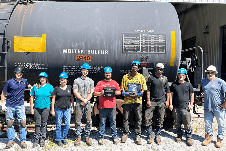 Group photo of employees in hard hats standing in front of a molten sulfur tank car, celebrating safety achievements with awards, showcasing teamwork and commitment to safety in the workplace.