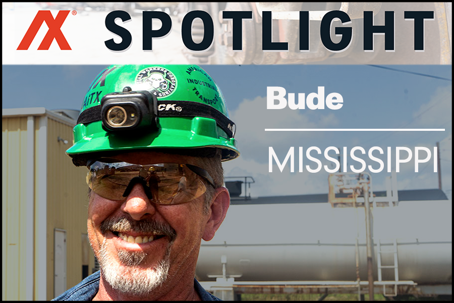 Smiling worker in a green hard hat with a headlamp, wearing safety glasses, in Bude, Mississippi. The background features industrial buildings and equipment, highlighting workplace safety and professionalism in the region.