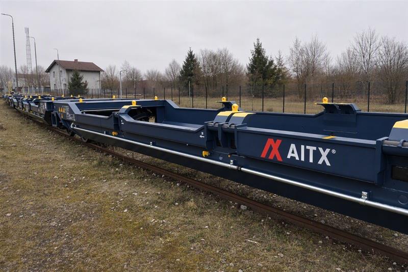 Blue transport containers on a railway track in Europe, featuring the AITX logo, with a backdrop of trees and a building in a cloudy environment.