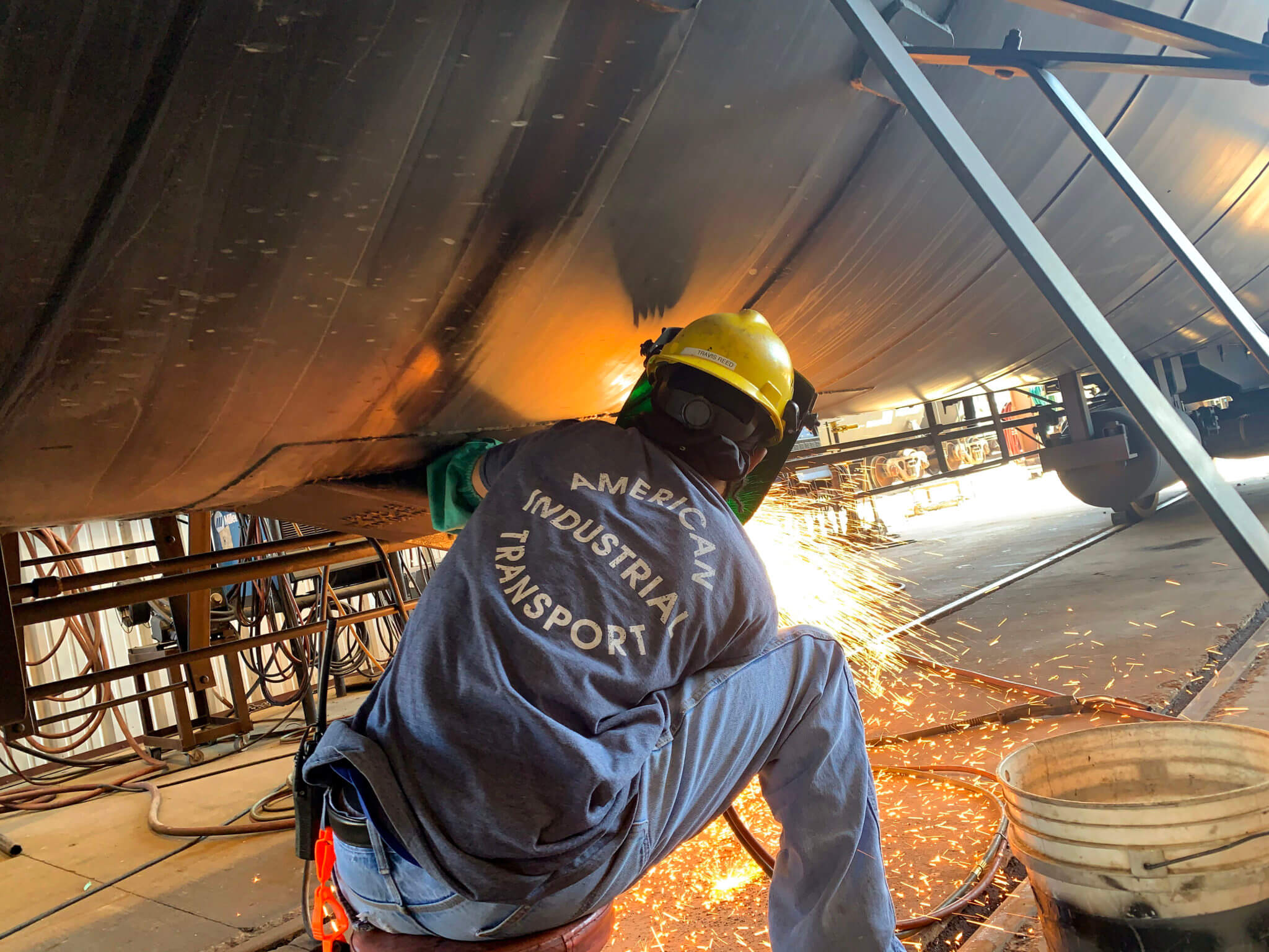 Welder working on the underside of a ship, creating sparks while performing metalwork in an industrial setting. The worker is wearing safety gear, including a helmet and gloves, highlighting the importance of safety in welding operations.