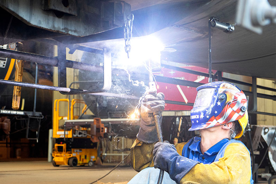Welder in protective gear using a welding torch to work on metal structures in a workshop, with sparks and smoke visible.