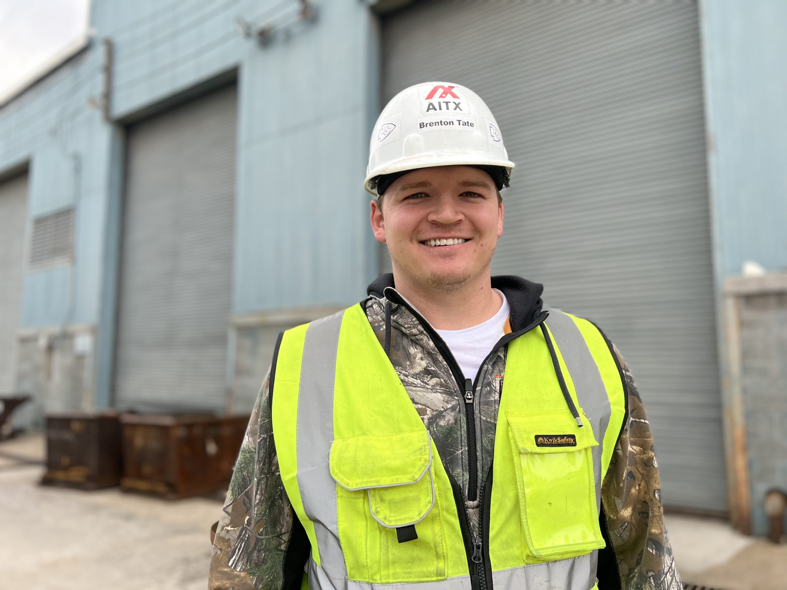 Portrait of a smiling construction worker wearing a hard hat and bright safety vest, standing in front of industrial buildings with metal containers in the background.