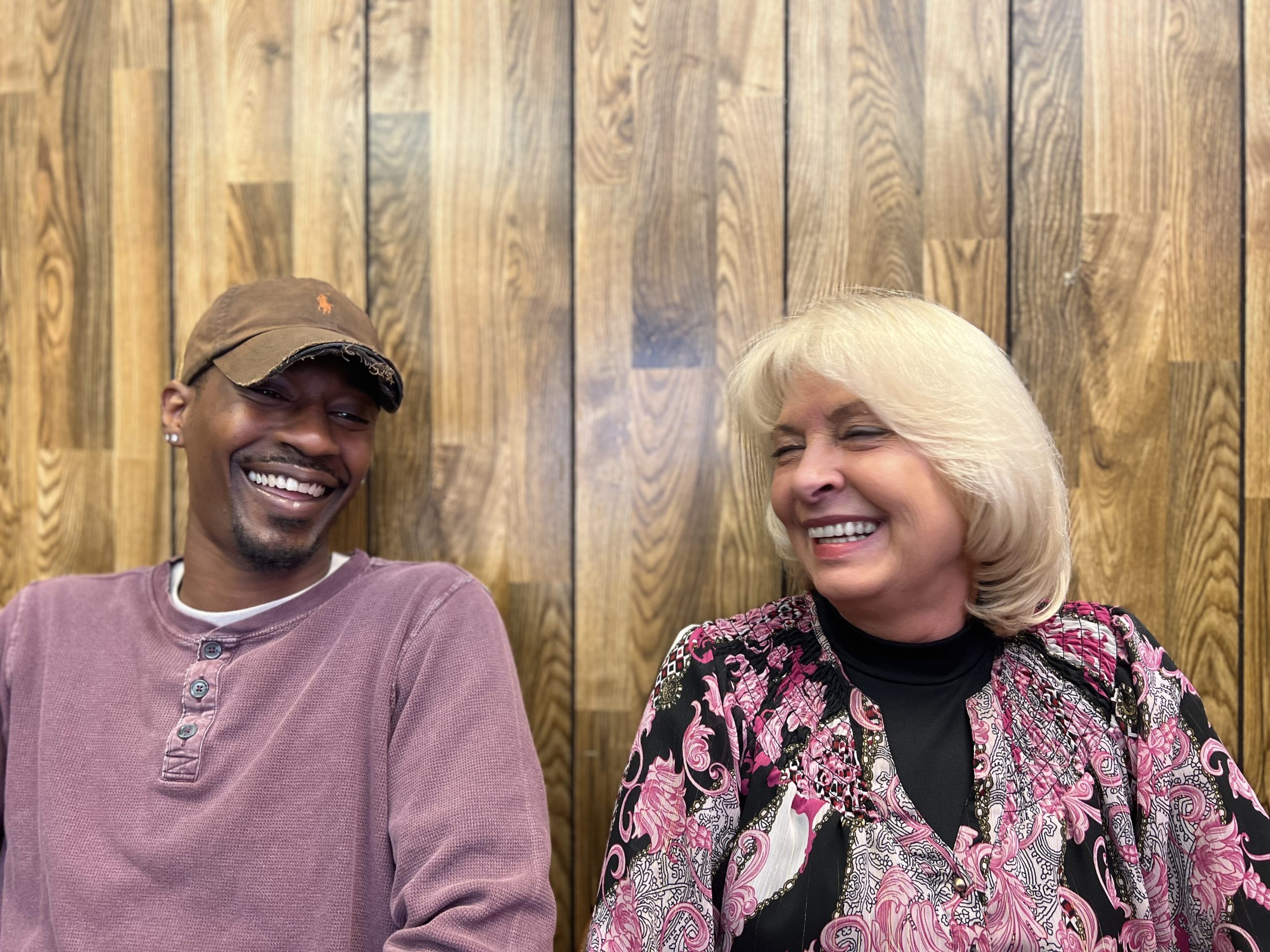 Two people, a man in a brown cap and a woman with blonde hair, are laughing together in front of a wooden wall. The scene captures a joyful moment, highlighting their friendly interaction and warm expressions.
