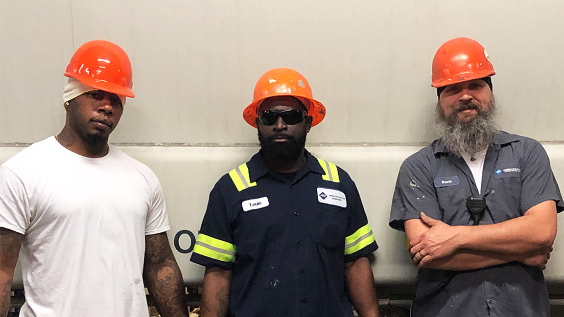 Three construction workers wearing orange hard hats and safety gear stand together in an industrial setting. The worker in the center has a black shirt with reflective stripes and sunglasses, while the others wear white and gray shirts. The background features a large, neutral-colored wall.