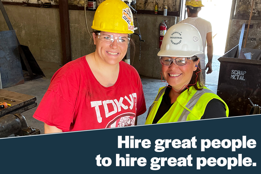 Two women in safety gear smile in a workshop, promoting teamwork and hiring skilled individuals in the industrial sector. The background features tools and equipment, emphasizing a hands-on work environment.