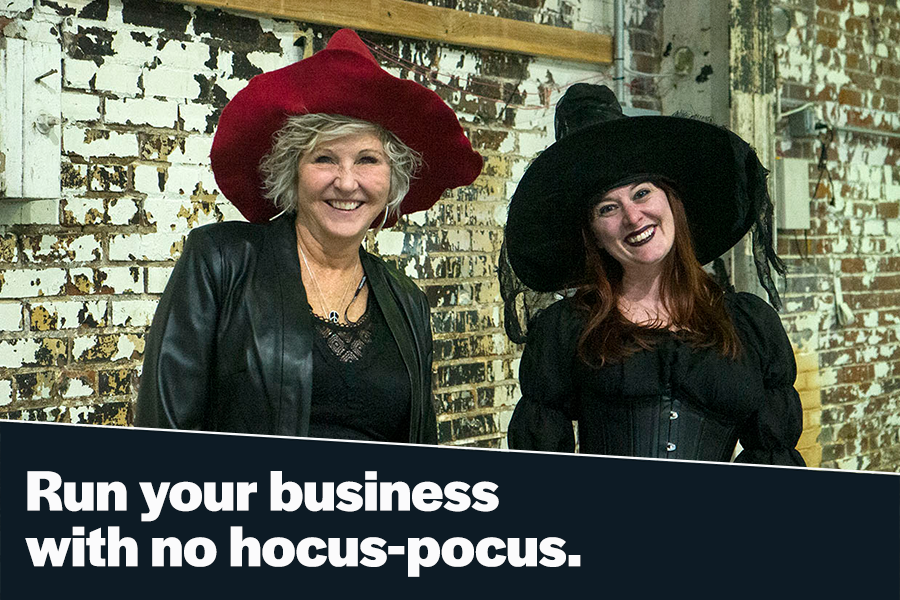 Two women wearing witch hats and costumes smile in front of a rustic brick wall, promoting a business theme with a playful tagline about running a business without tricks.