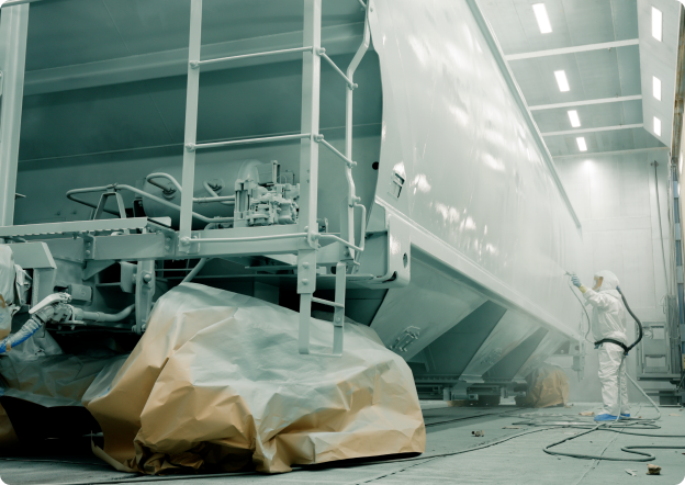 Industrial worker spray painting a large cargo container in a well-lit spray booth, with protective coverings on the floor and equipment for safety.