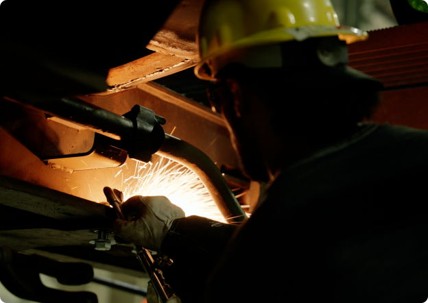 A worker wearing a hard hat and gloves performs welding on metal equipment, with sparks flying in a dimly lit workshop environment.