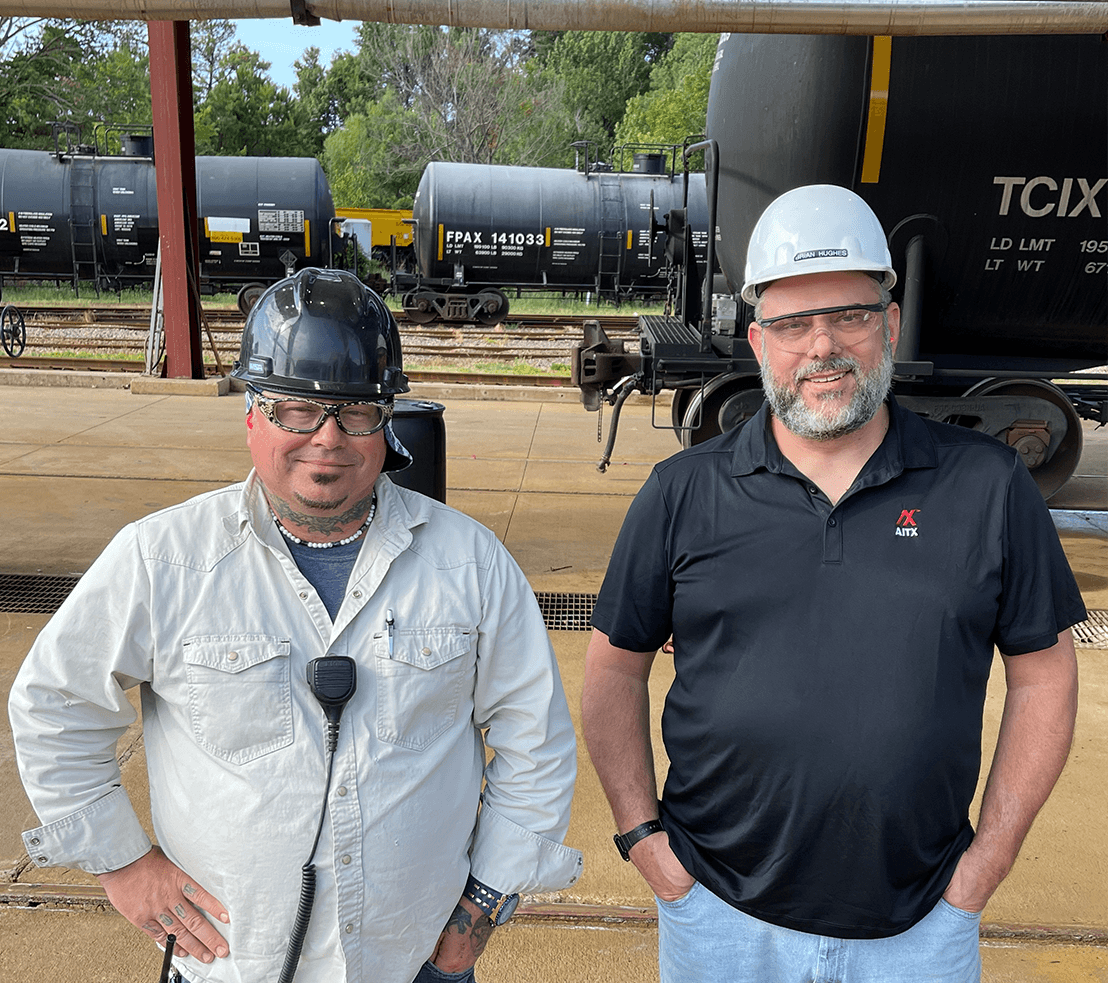 Two men in safety gear, including hard hats and safety glasses, stand in front of a railway yard with black tank cars in the background. The man on the left wears a light-colored shirt and a communication device, while the man on the right is in a dark polo shirt with a company logo. This image highlights safety practices in the industrial sector.