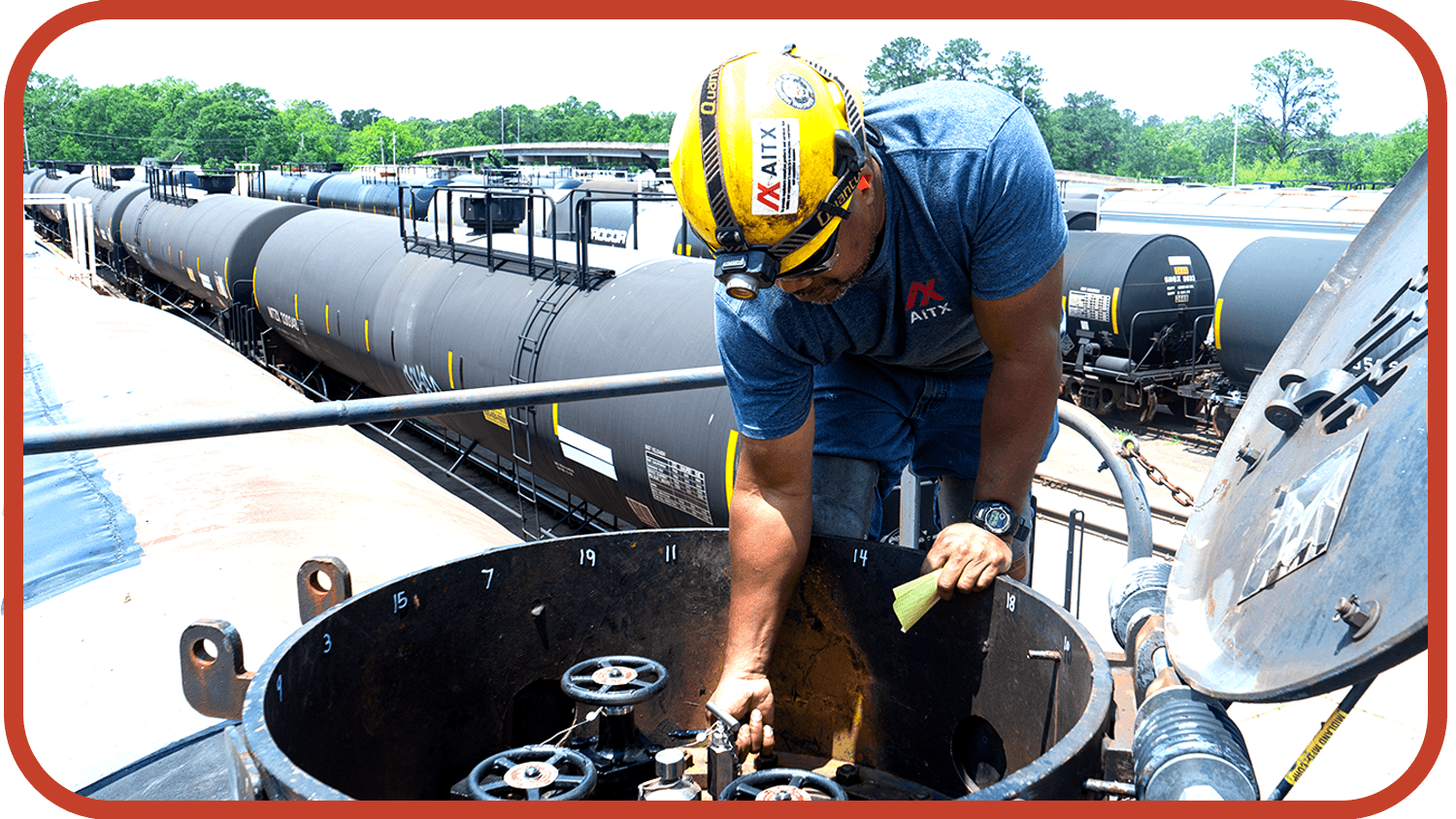 Worker inspecting a tank car at a rail yard, using tools and wearing safety gear, with multiple tank cars in the background under clear blue skies.