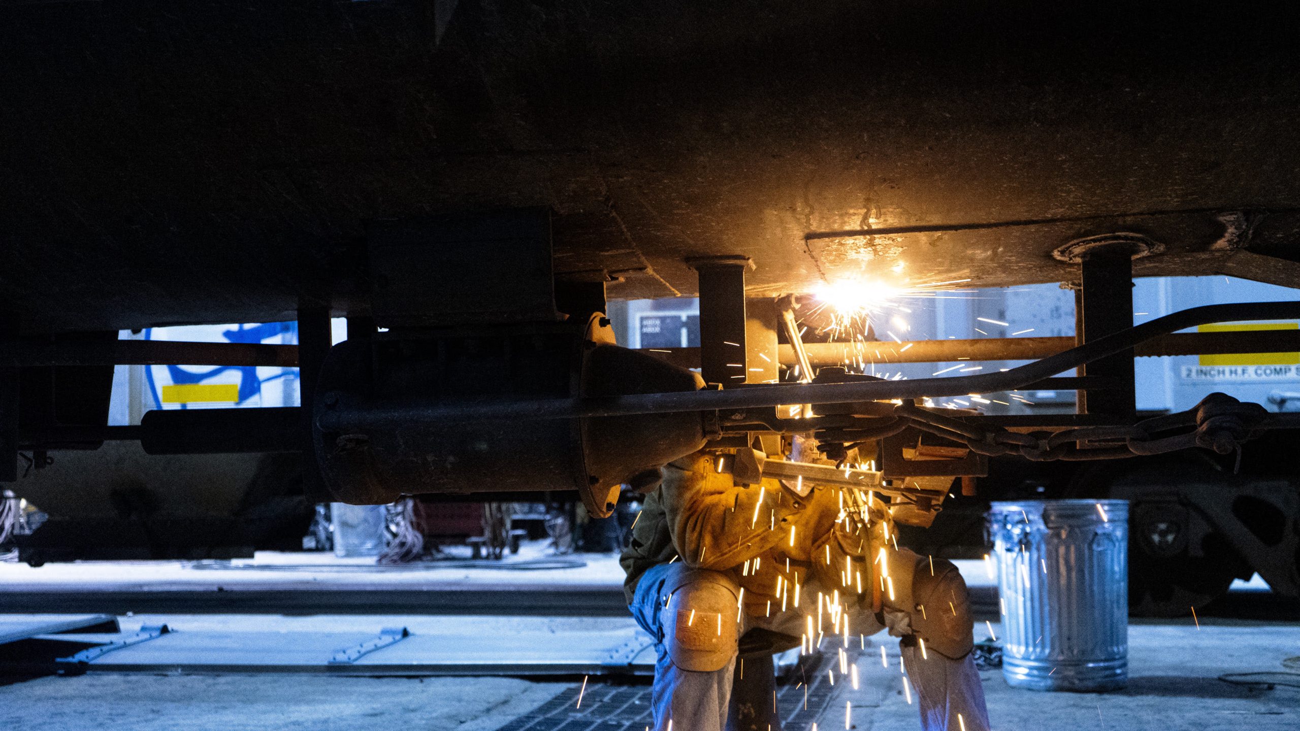 Welder performing maintenance on heavy machinery, sparks flying from welding torch in a dimly lit workshop.