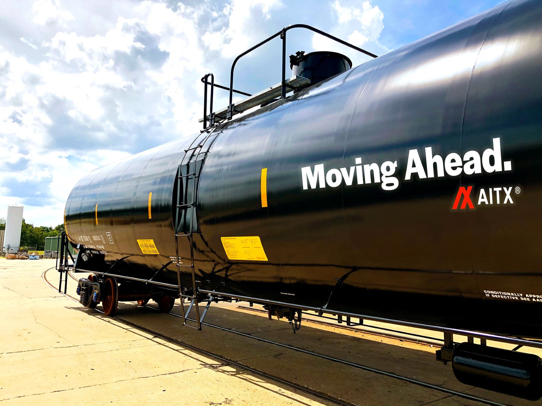 Black tank car on railway tracks with the text "Moving Ahead" and the logo of AITX, set against a partly cloudy sky. The image showcases industrial transportation and innovation in logistics.