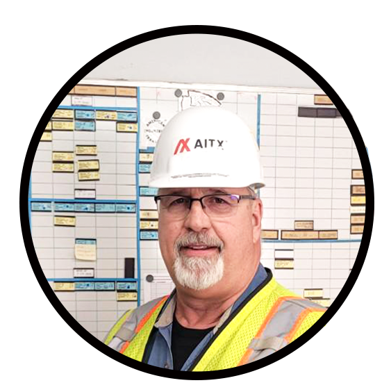 Construction professional wearing a hard hat and safety vest, standing in front of a project management board filled with schedules and tasks.