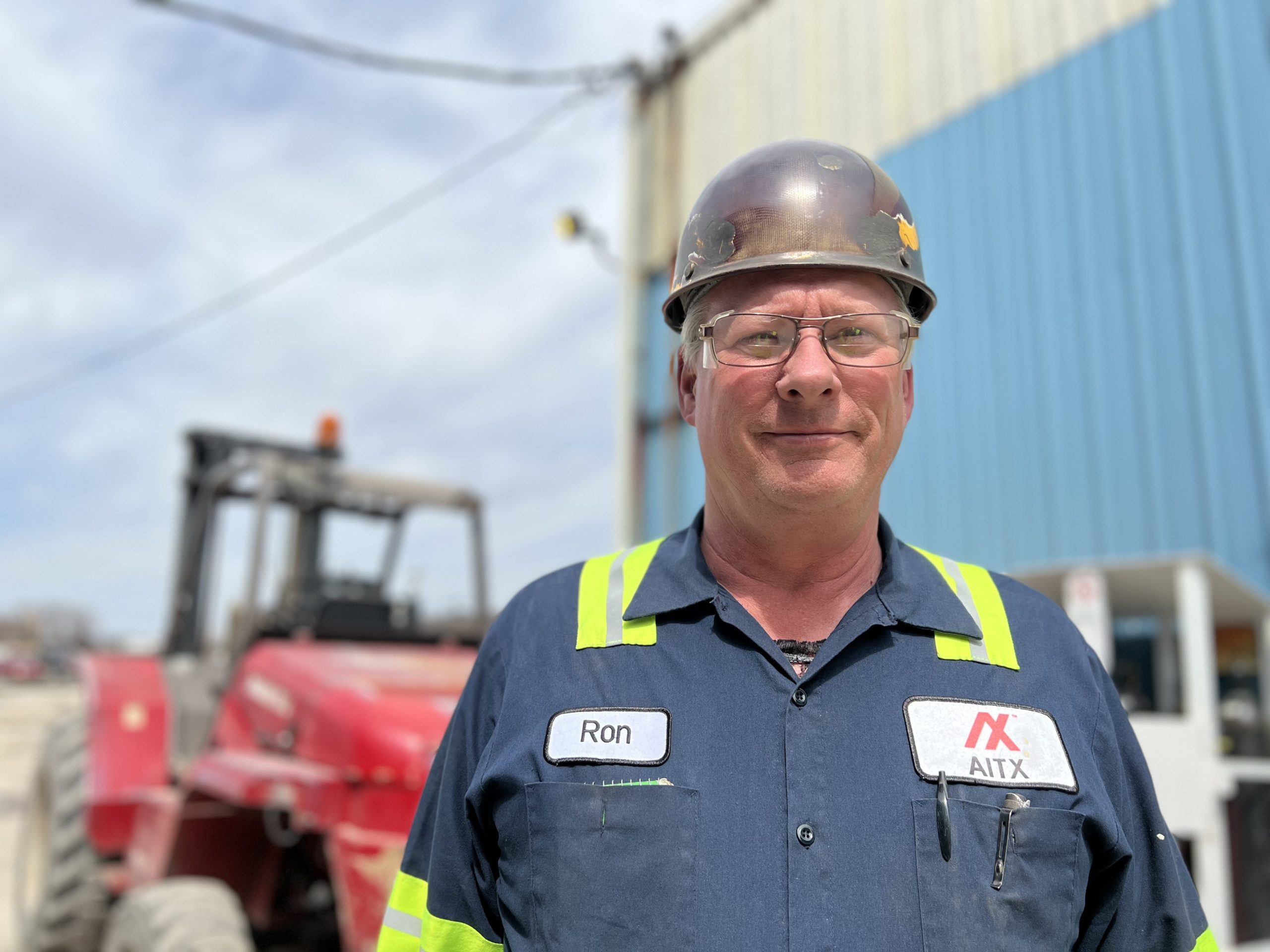 Construction worker wearing a hard hat and safety glasses, standing in front of a red tractor in an industrial setting, showcasing a professional demeanor and safety gear.