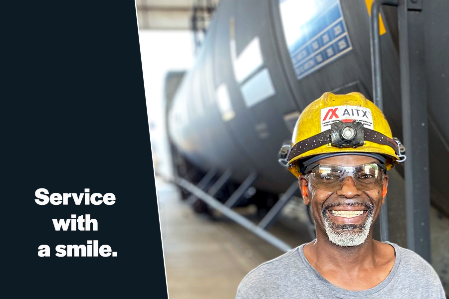 Smiling worker in safety gear, including a hard hat and goggles, standing in front of industrial tanks, showcasing a positive attitude in a professional environment.