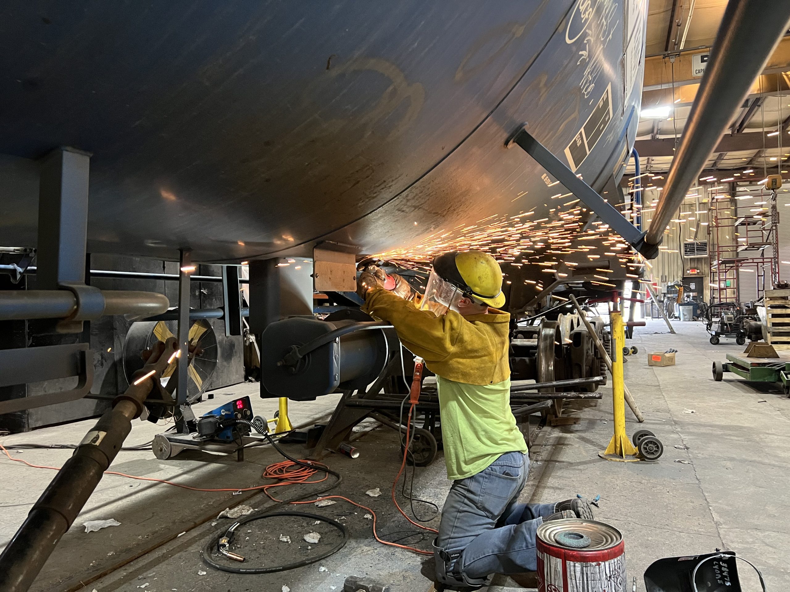 Welder working on the underside of a large metal structure in a workshop, sparks flying from the welding process, with tools and equipment visible in the background.