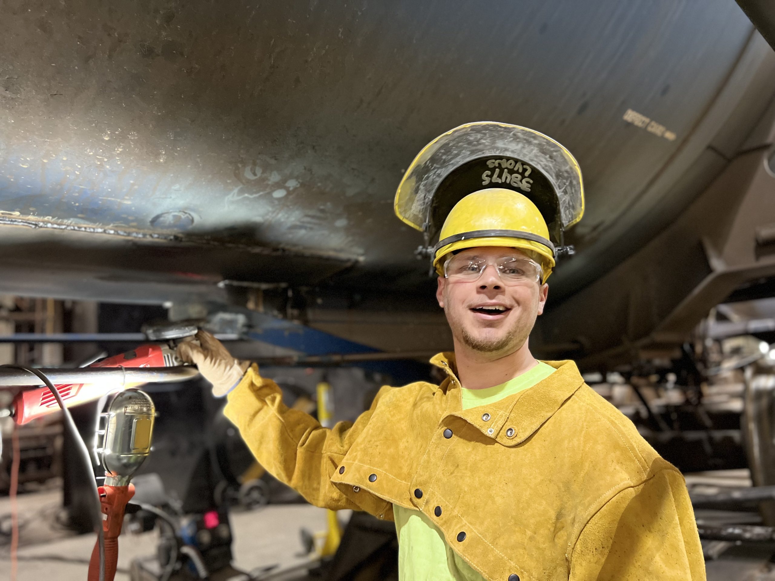 Young male welder wearing protective gear, including a yellow hard hat and gloves, smiles while working on a metal structure with a welding tool in a workshop setting. The scene highlights safety measures and the welding process in an industrial environment.