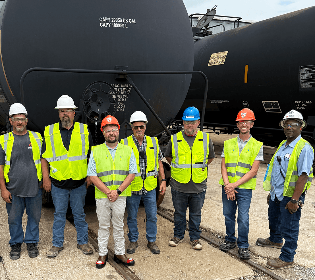 Group of seven workers in safety vests and hard hats posing in front of a large black tank car, showcasing teamwork and safety in the industrial environment.