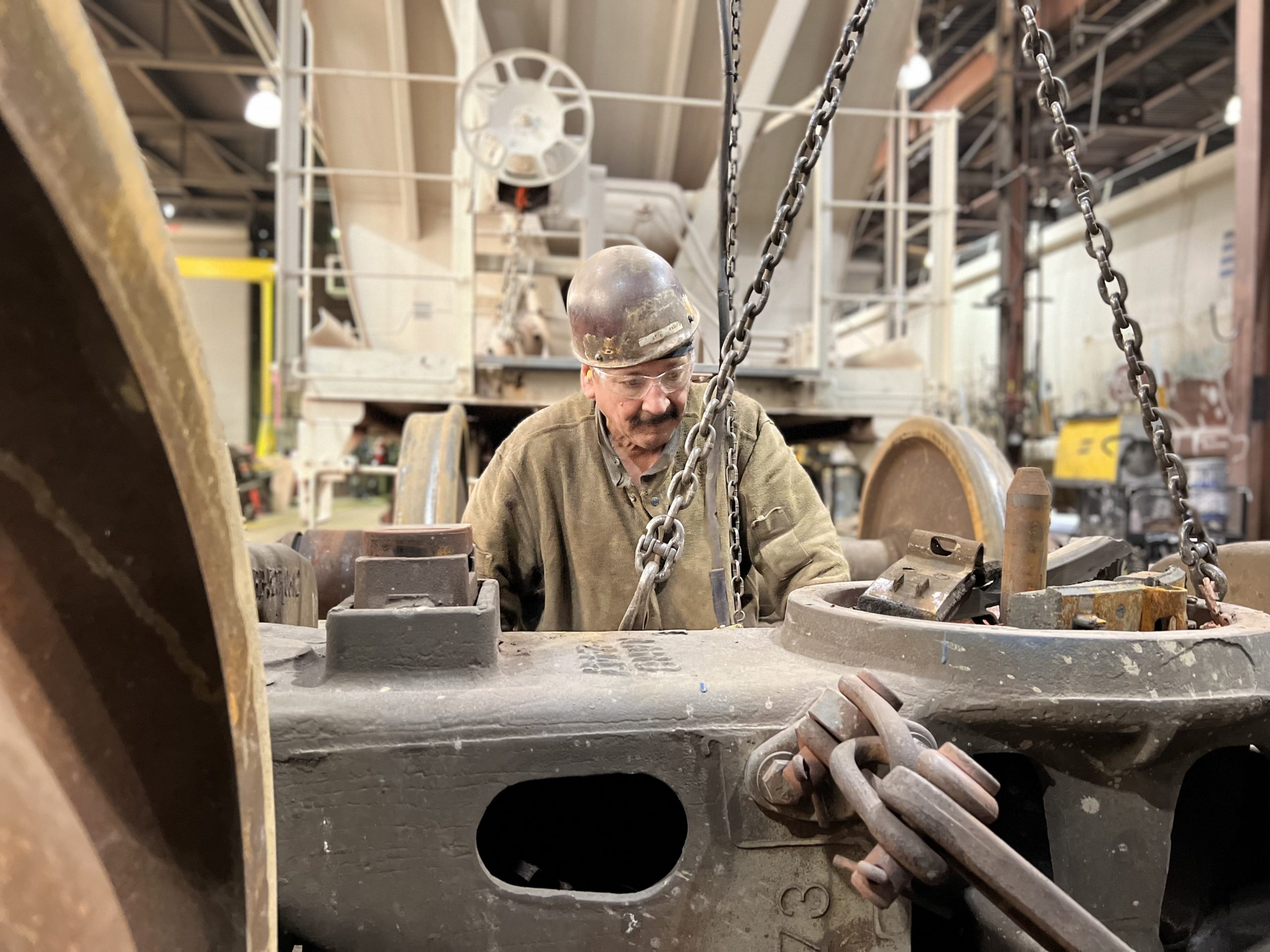 Worker in a workshop inspecting and repairing heavy machinery, with a focus on mechanical components and equipment in the background. The image captures the industrial setting and the detailed work involved in maintenance tasks.